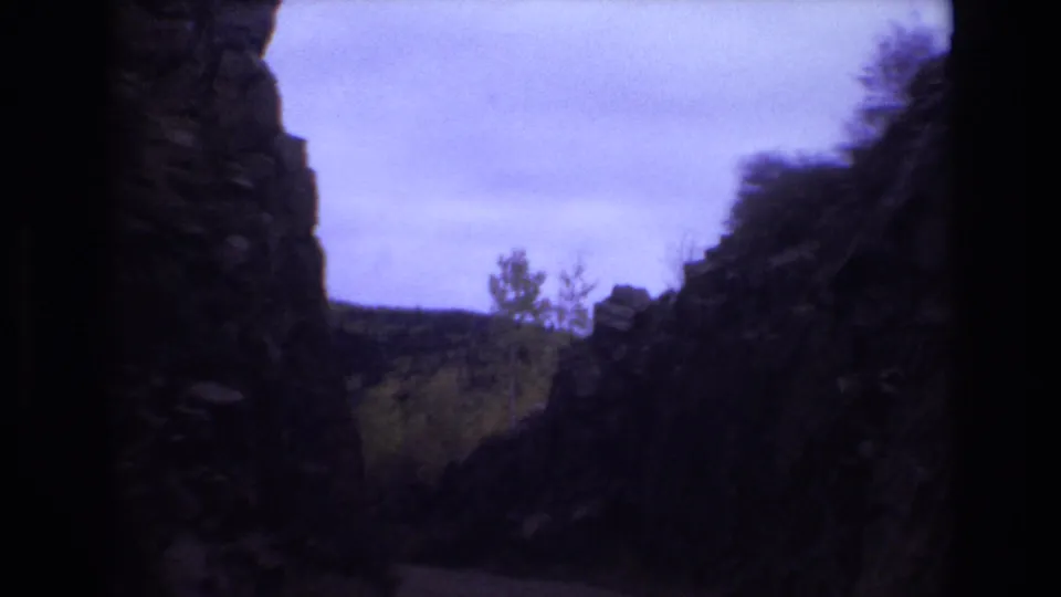 1973 Colorado Springs home-movie frame showing a narrow rocky passage, small trees, distant hillside, and purple-tinted sky as the camera continues forward.
