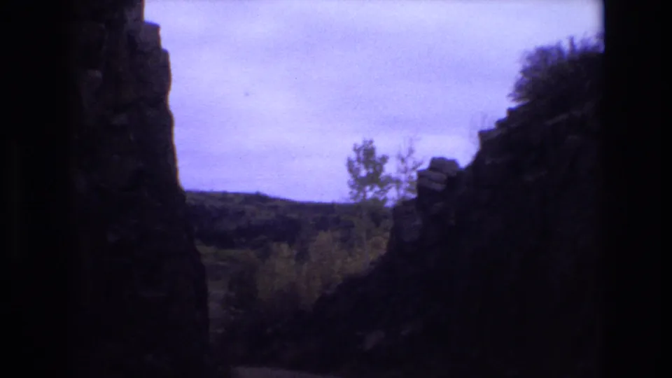 1973 Colorado Springs 8mm frame showing shadowed rock formations around a narrow view of wooded hills and a cloudy purple-tinged sky.