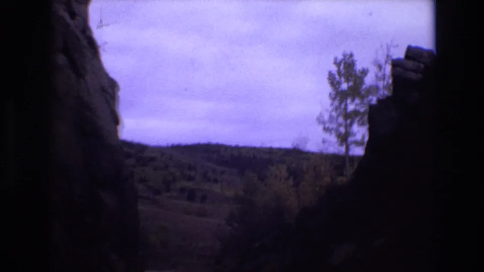 1973 Colorado Springs 8mm frame with a dark vertical rock formation dividing the view toward rolling wooded terrain and a cloudy sky.