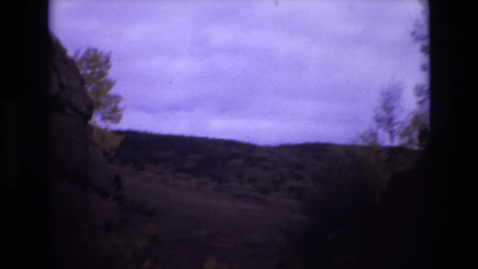 1973 Colorado Springs 8mm frame looking past a shadowed rock face toward trees, brush, low hills, and a cloudy sky.