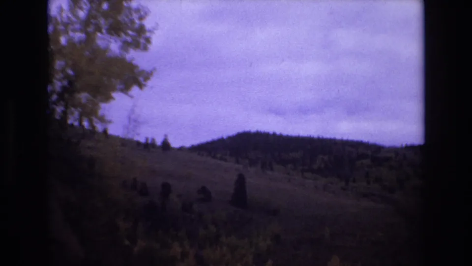 1973 Colorado Springs 8mm frame showing a distant hillside with trees and brush, framed by dark rocky edges and a purple-blue cloudy sky.