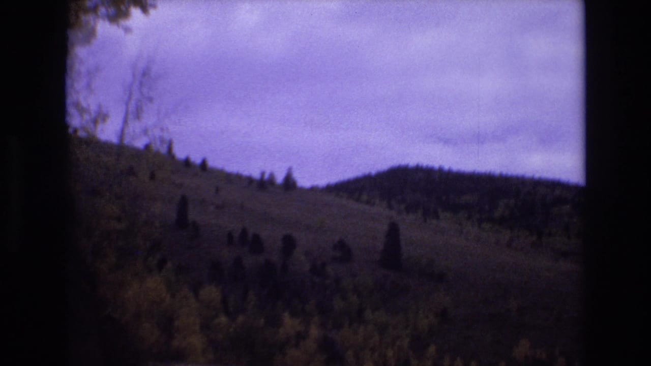 1973 Colorado Springs home-movie frame showing a rolling hillside with scattered evergreens under a cloudy sky, seen through dark 8mm film borders.