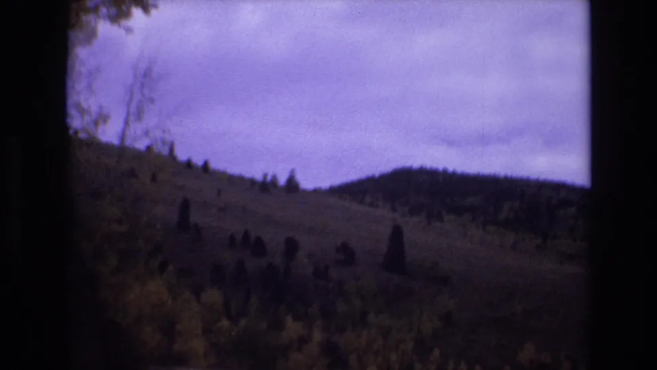 1973 Colorado Springs home-movie frame showing a rolling hillside with scattered evergreens under a cloudy sky, seen through dark 8mm film borders.