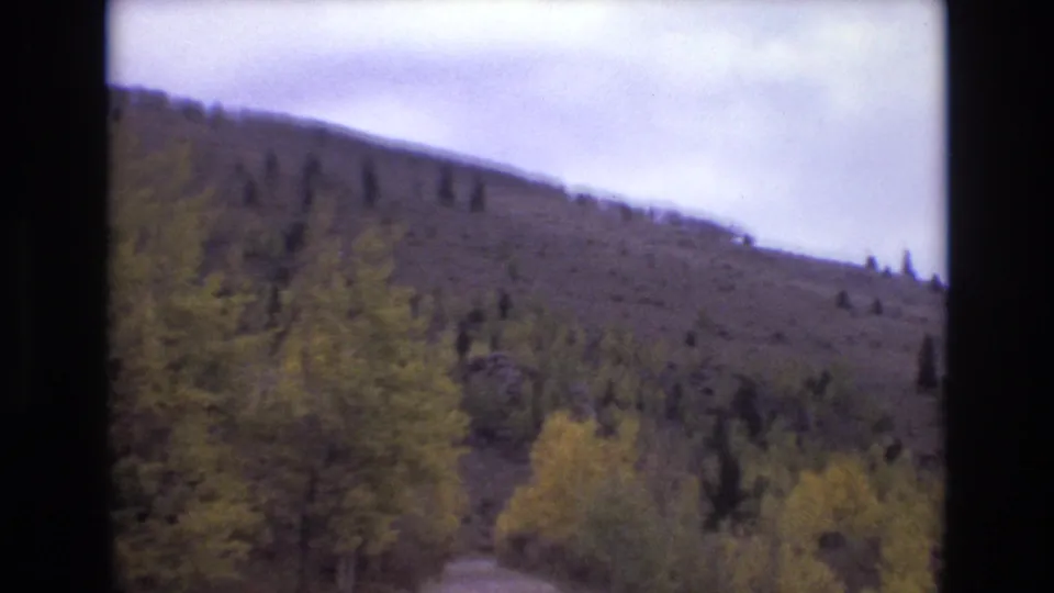 1973 Colorado Springs home-movie frame showing yellowing roadside foliage in front of a tree-dotted hillside and cloudy sky.