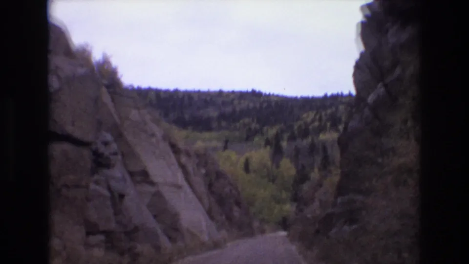 1973 Colorado mountain-road frame with dark film borders, rocky canyon walls, and a distant tree-covered slope ahead.