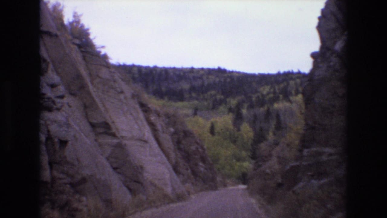 1973 8mm view in Colorado Springs showing a road entering a narrow rocky pass with evergreen forest and yellow-green foliage beyond.