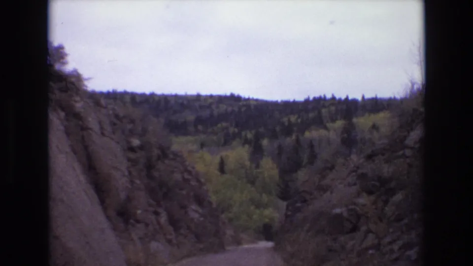 8mm 1973 Colorado Springs travel frame with the road dropping through a narrow rocky passage toward mixed evergreen and yellow foliage.