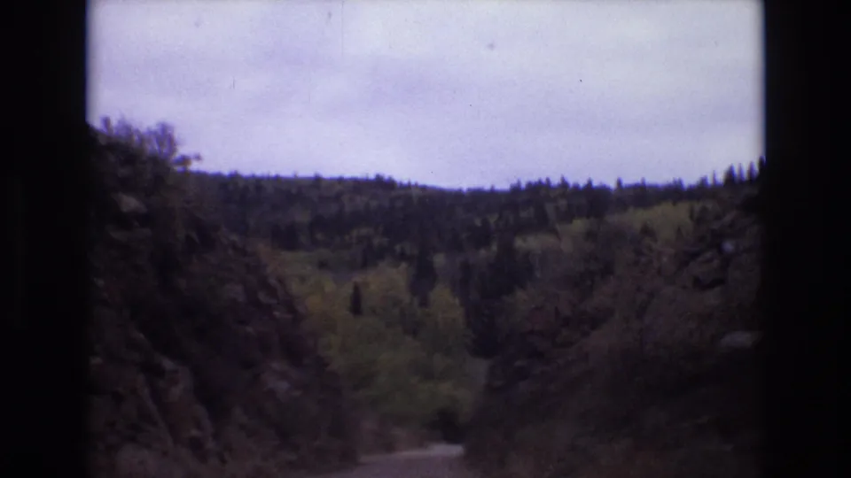 Faded 8mm frame from Colorado Springs in 1973 showing a roadway between dark rocky edges and a forested hillside under an overcast sky.
