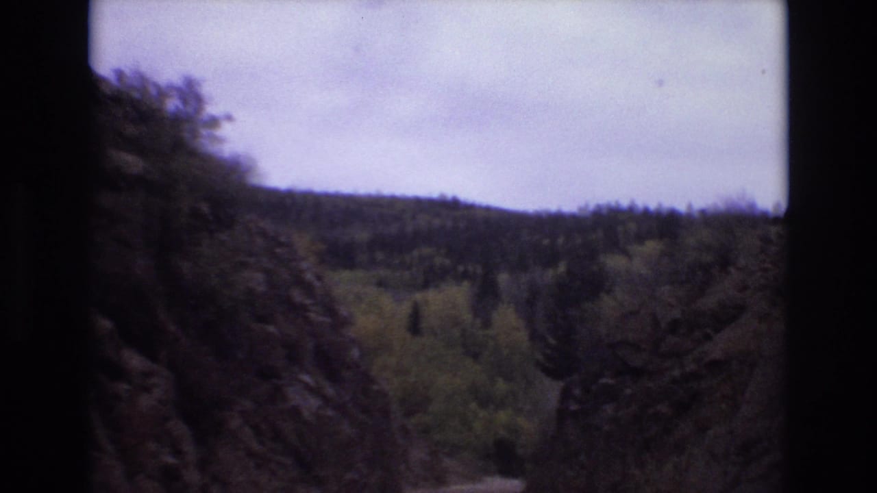 1973 Colorado Springs home-movie frame showing a forward road view between dark rocky cliffs, trees, distant wooded hills, and a pale overcast sky.