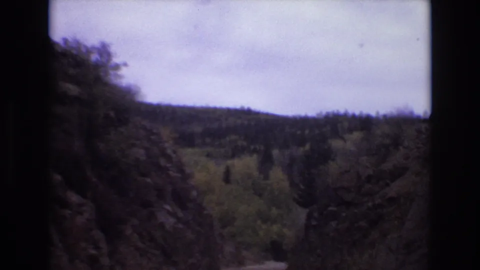 1973 Colorado Springs home-movie frame showing a forward road view between dark rocky cliffs, trees, distant wooded hills, and a pale overcast sky.