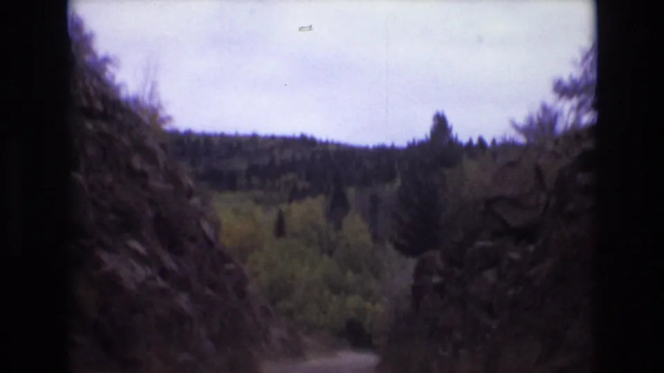 Colorado Springs 1973 travel frame looking ahead through a narrow rocky pass with evergreen trees, yellow-green foliage, and a low wooded ridge beyond.
