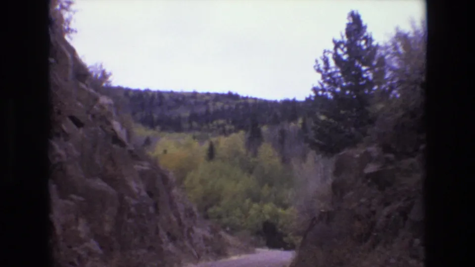 Forward-facing 8mm frame from the Colorado Springs road trip showing a curving dirt or gravel road below steep rock, pine trees, and early autumn foliage.