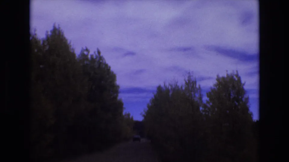 1973 home-movie frame from a moving vehicle showing a narrow wooded road, dark roadside trees, and a small distant vehicle shape under the blue evening sky.