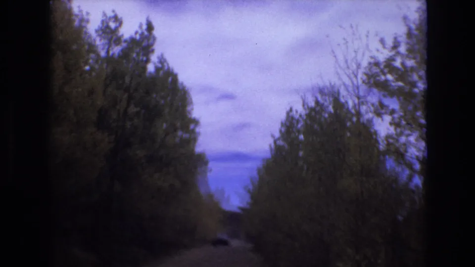Low-light 8mm frame of a Colorado Springs-area rural road in 1973, with trees framing the roadway and dusk clouds across a saturated blue-purple sky.