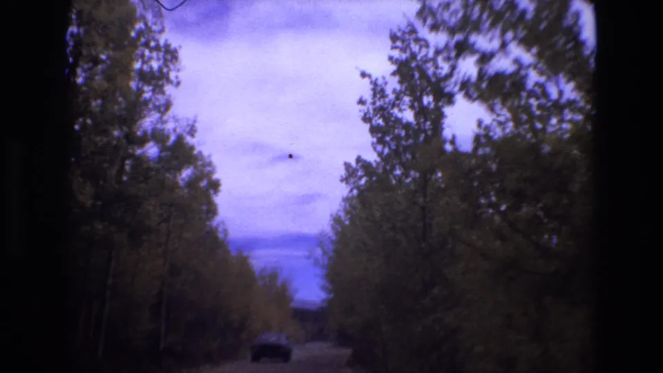 A moving 8mm view along a wooded dirt road near Colorado Springs, with dense trees, muted sky, and a dark car ahead in the lane.
