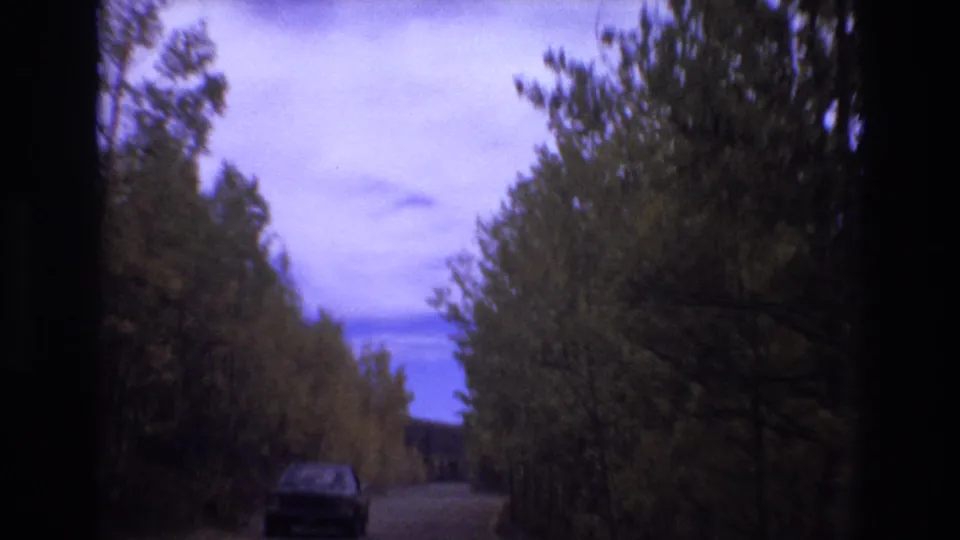 The 1973 home-movie frame shows the vehicle ahead closer on the unpaved road, framed by tall green and yellow trees and 8mm edge shadows.