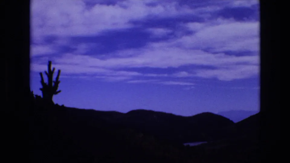 1973 Colorado Springs home-movie frame with a cactus-like plant silhouette at left, dark rolling hills, and a saturated blue sky with soft clouds.