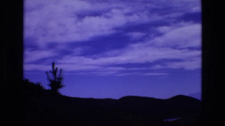 1970s 8mm landscape frame showing a dark sloping foreground, cactus-like silhouette, low hills, and a blue-purple clouded sky with soft archival grain.