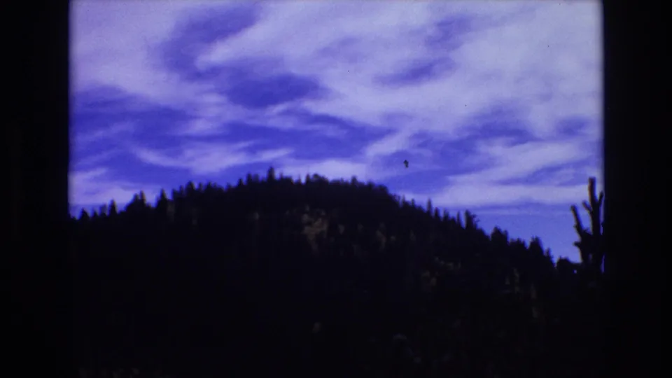 1973 8mm Colorado Springs landscape frame showing a dark ridgeline, scattered tree silhouettes, and swirling blue-purple clouds overhead.