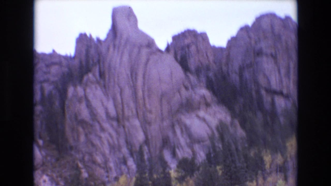 1973 Colorado Springs 8mm frame with a pointed rock outcrop, ridged cliff faces, evergreens, and fall foliage beneath a bright sky.