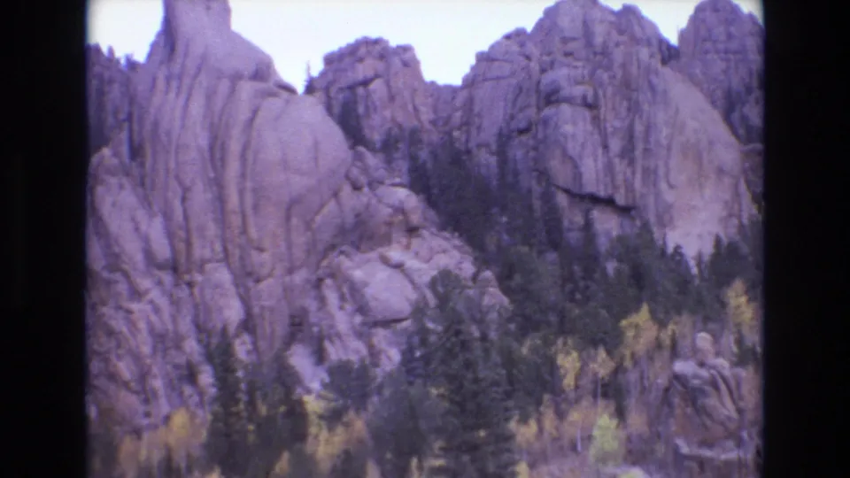 1973 8mm home-movie frame from Colorado Springs showing steep textured rock faces, dark pines, and patches of yellow autumn leaves.