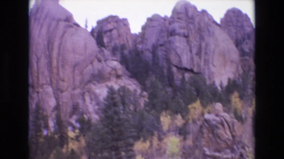 1973 Colorado Springs 8mm frame with a rocky mountainside, clustered evergreen pines, and small patches of yellow foliage beneath an overcast-looking sky.