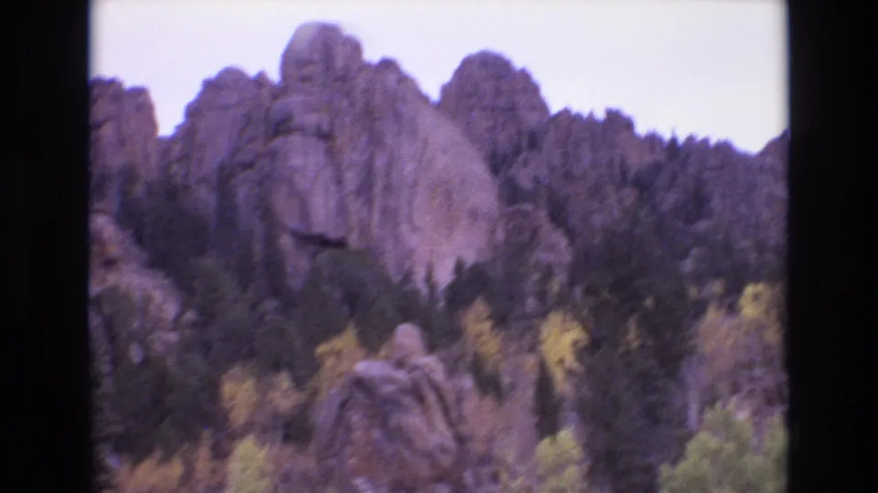 1973 Colorado Springs 8mm frame showing a pine-covered slope beneath rounded rock formations, with yellow autumn trees and a large boulder in the foreground.