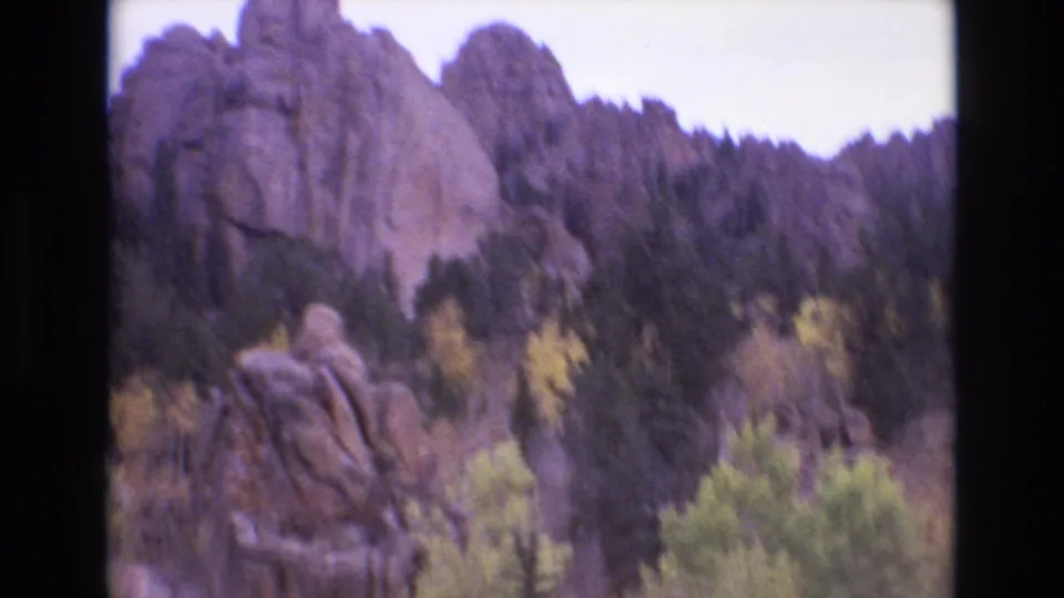1973 Colorado Springs 8mm frame looking across a forested slope toward a rounded cliff face, with evergreen pines and yellow-green trees in the lower frame.