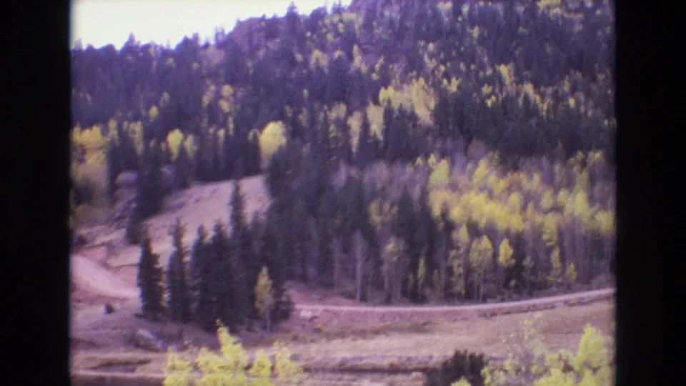 1973 Colorado Springs 8mm frame showing a winding dirt road at the base of a forested mountain with evergreens and yellow autumn trees.