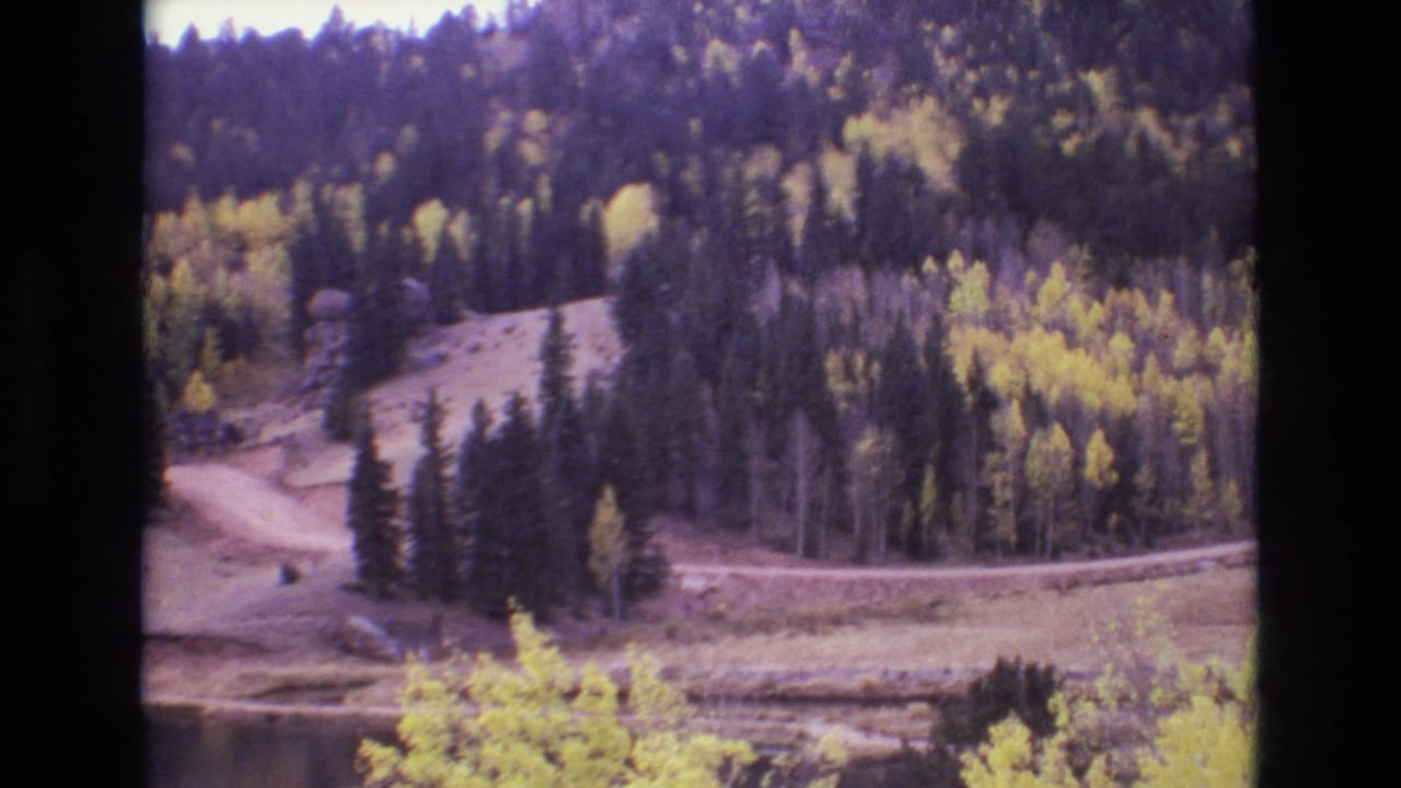 1973 Colorado Springs-area home-movie frame showing a wooded mountain slope with yellow aspens, evergreen pines, a winding dirt road, and the edge of a calm lake.