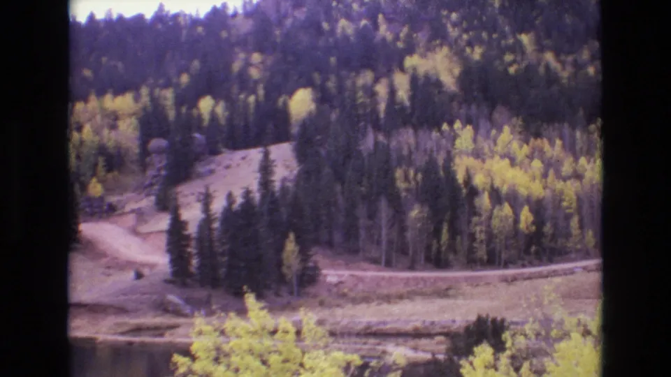 1973 Colorado Springs-area home-movie frame showing a wooded mountain slope with yellow aspens, evergreen pines, a winding dirt road, and the edge of a calm lake.