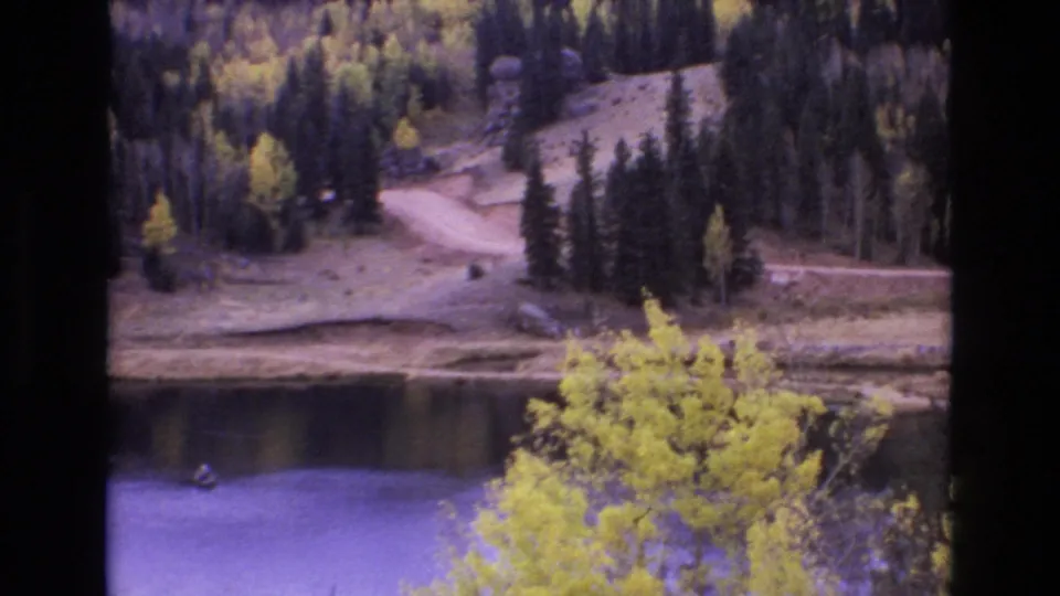 1973 Colorado Springs-area 8mm frame showing calm lake water, a wooded shoreline, yellow aspens, pine trees, and a winding dirt road across the hillside.