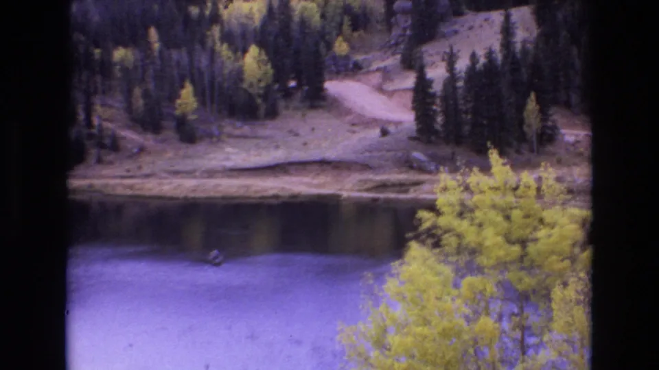 1973 Colorado Springs-area home-movie frame with a wider view of the lake, yellow aspens in the foreground, pine-covered slopes, a dirt road, and a small boat-like shape on the water.