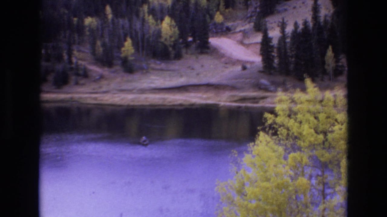 1973 Colorado Springs home-movie frame showing a distant lone fisherman in a small dinghy on calm water, with yellow trees, evergreens, and a pale shoreline beyond.