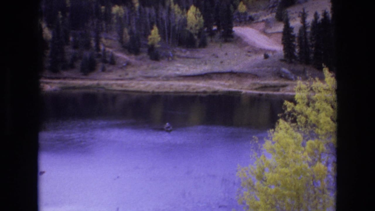 1973 Colorado Springs 8mm frame with a lone fisherman in a small boat near the far shoreline, framed by purple-blue water, yellow foliage, and dark evergreens.