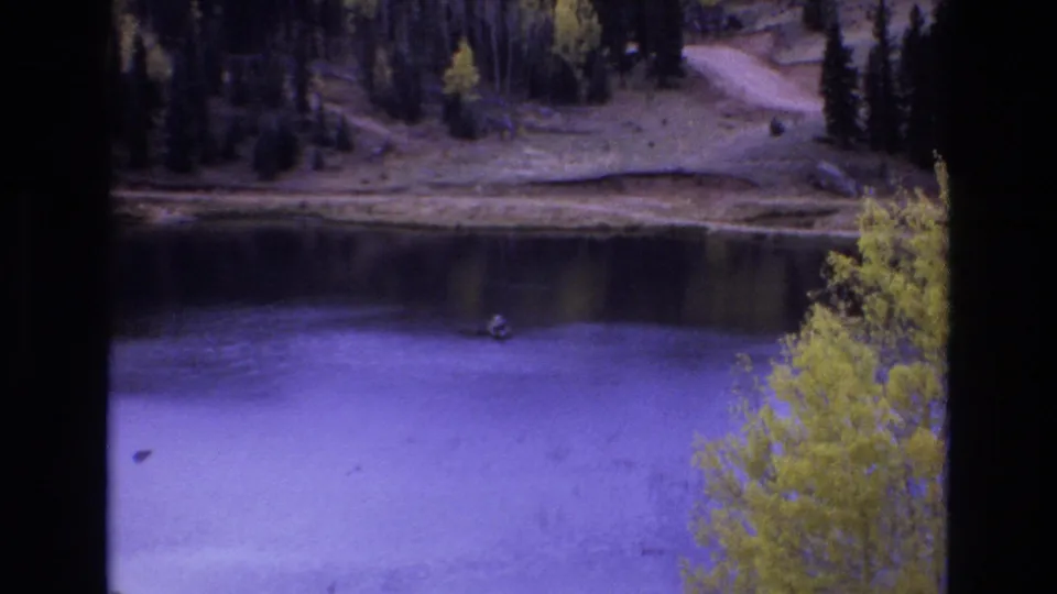 Home-movie frame from 1973 Colorado Springs showing a distant fisherman seated in a dinghy on a calm lake, with autumn trees and wooded shore visible beyond.