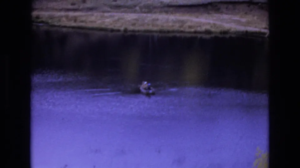 1973 Colorado Springs 8mm frame with a closer view of a lone fisherman in a dinghy on dark blue-purple water, with ripples spreading behind the boat.