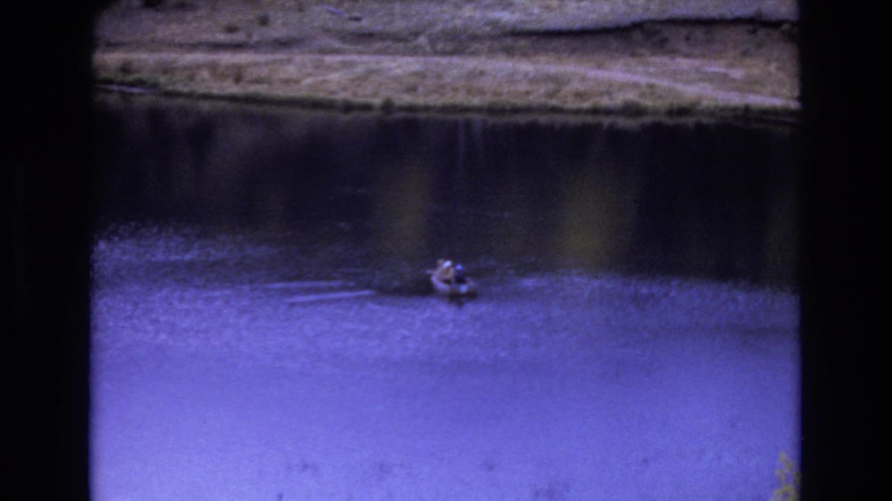 Small boat or skiff floating on a calm lake in 1973 Colorado Springs home-movie footage, with ripples spreading across blue-tinted water near a shadowed bank.