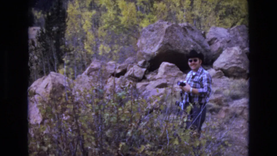 Adult man in a plaid shirt, sunglasses, and brimmed western-style hat holds a camera among large rocks and autumn brush in 1973 Colorado Springs home-movie footage.
