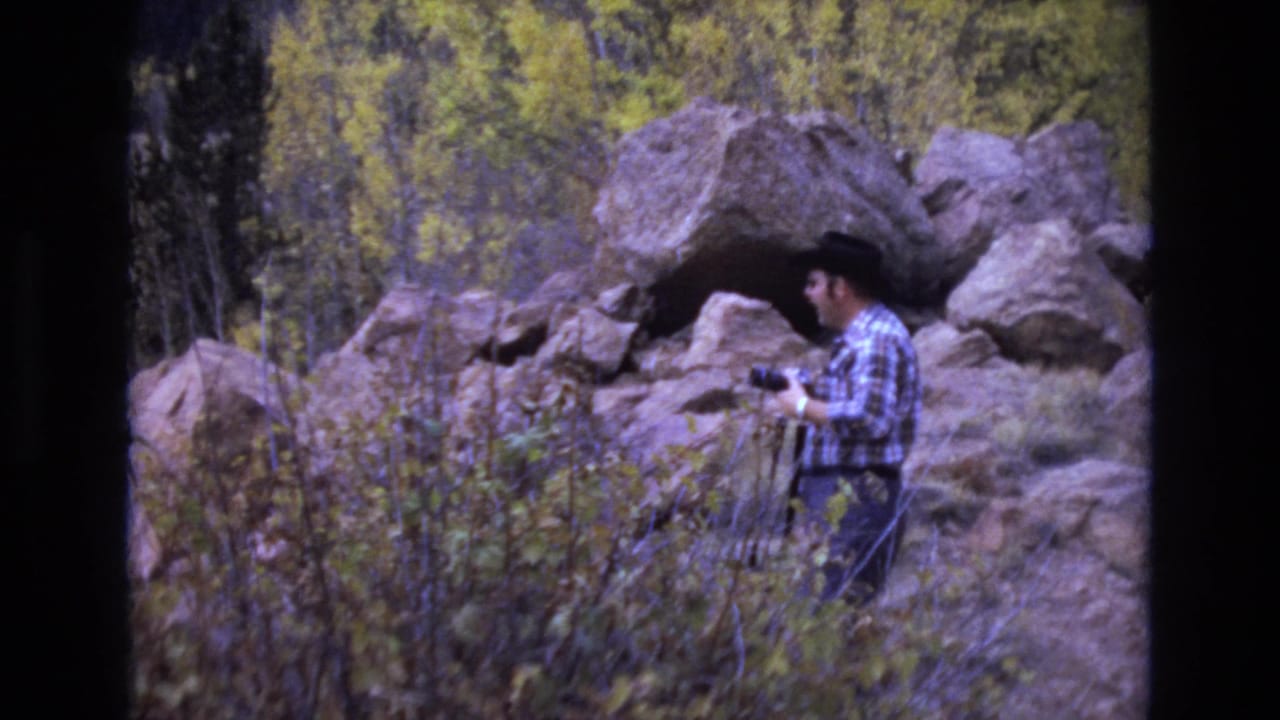In metadata-supported 1973 Colorado Springs footage, a man in a plaid shirt and dark cowboy hat stands near boulders and autumn brush while holding a camera.