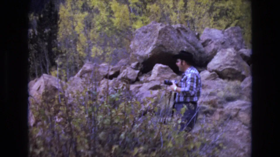 In metadata-supported 1973 Colorado Springs footage, a man in a plaid shirt and dark cowboy hat stands near boulders and autumn brush while holding a camera.