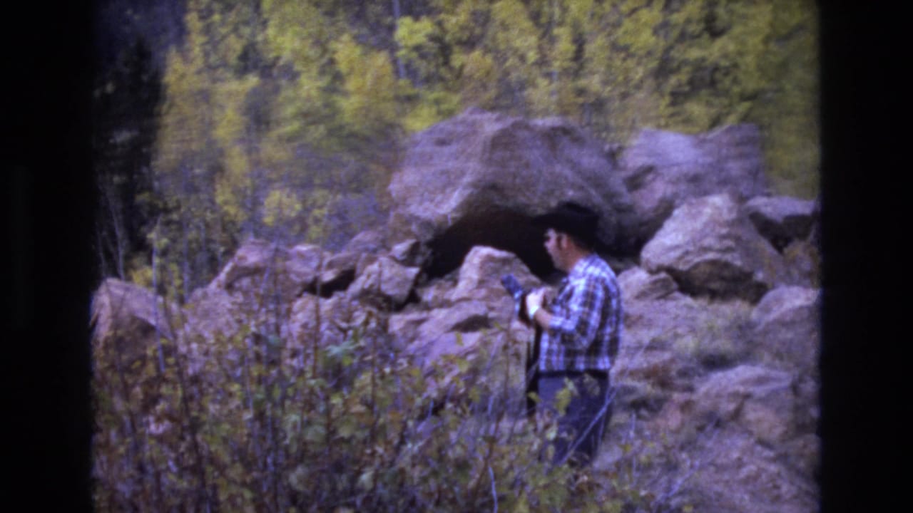 Man looks down at a camera while standing beside large rocks and shrubs.