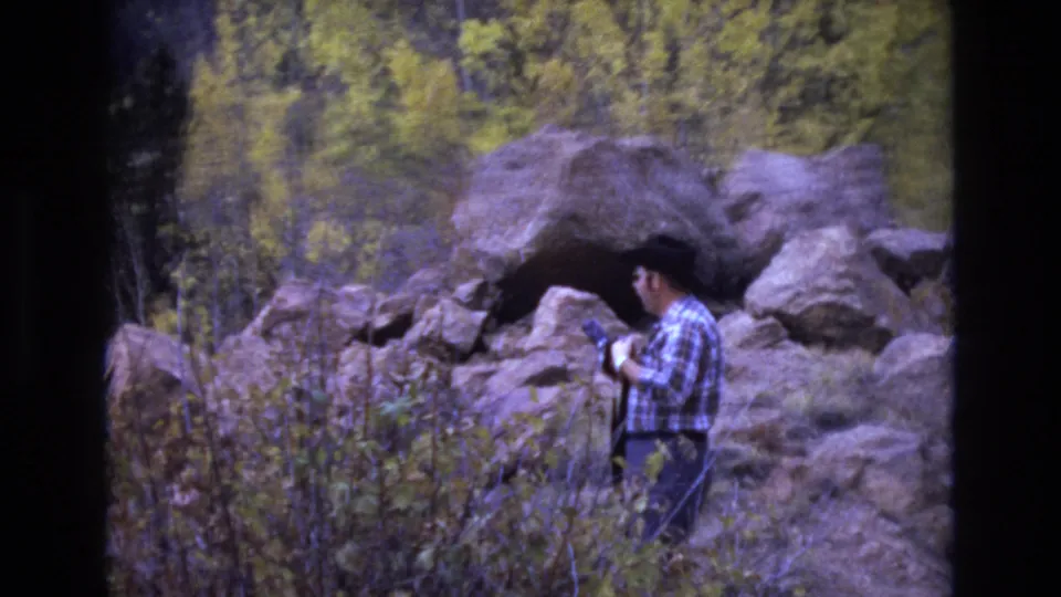 The man appears to check or handle his camera beside a cave-like recess in large boulders, with autumn foliage behind him in the Colorado Springs area.
