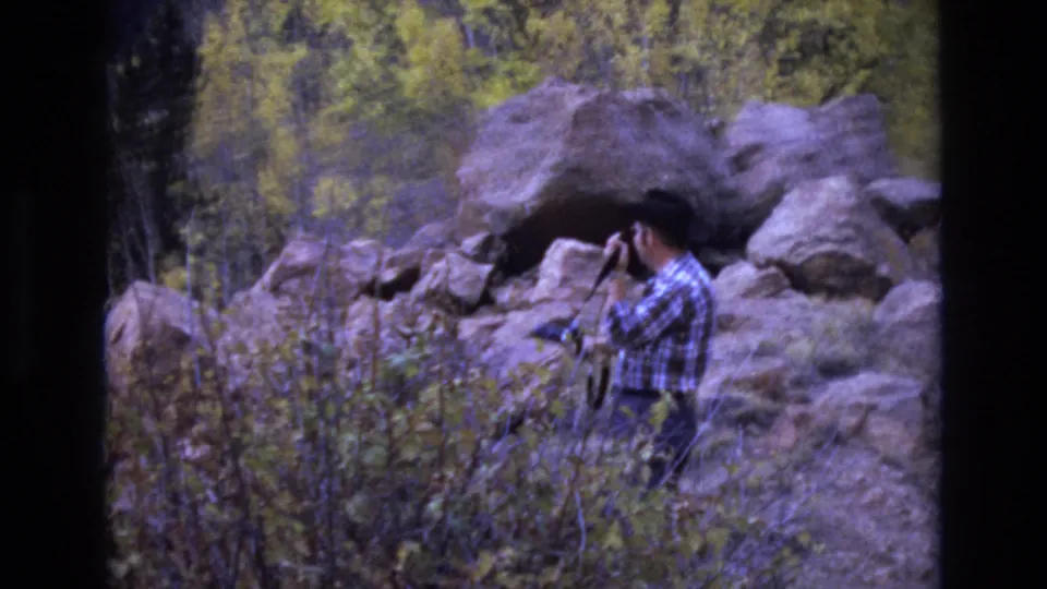 A man in a plaid shirt raises a camera to photograph or inspect a rocky cave-like opening, with brush and autumn trees filling the frame.