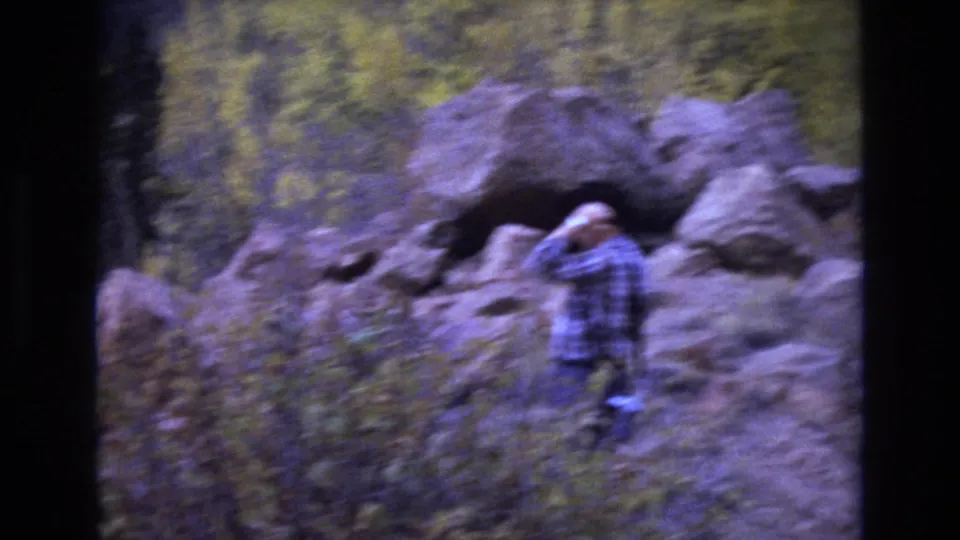 A softer handheld frame shows the man near the boulders, lifting an arm close to his hat or head as he stands among brush in the autumn landscape.