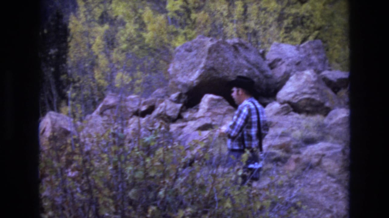 In Colorado Springs home-movie footage from 1973, a man in a plaid shirt and dark brimmed hat stands near boulders and autumn brush.