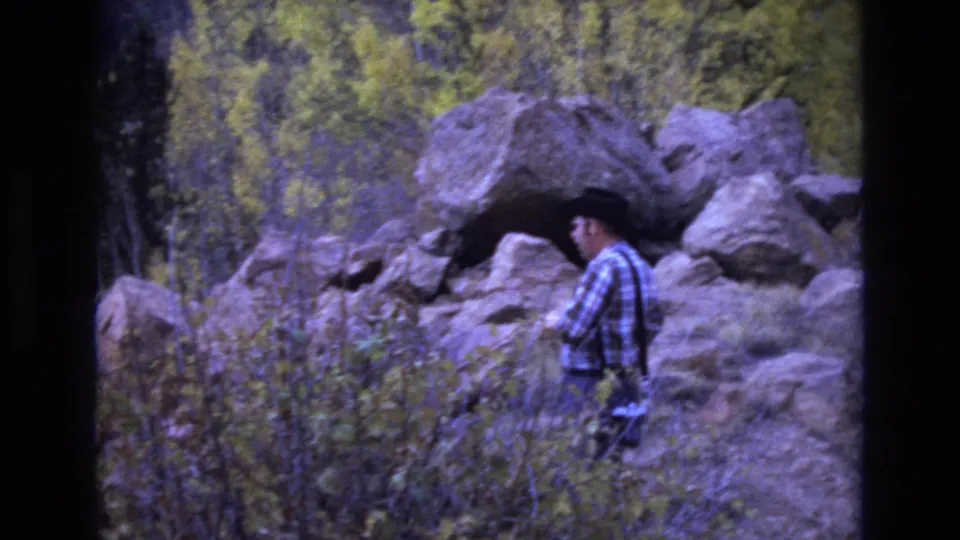 In Colorado Springs home-movie footage from 1973, a man in a plaid shirt and dark brimmed hat stands near boulders and autumn brush.