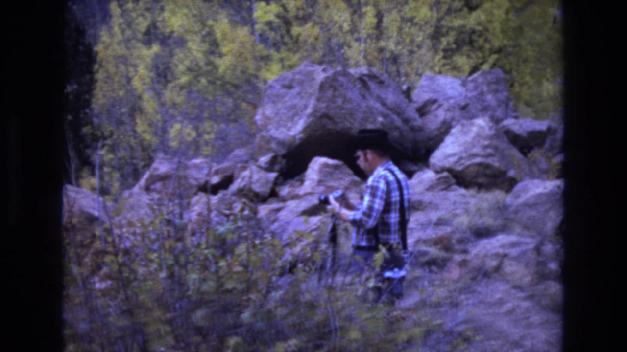 Man looks down while holding or checking a camera near boulders and brush.