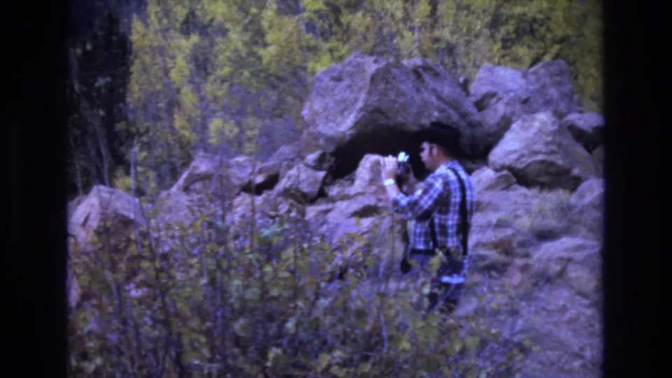 The man raises a camera toward the rocky wooded landscape, with large boulders and yellow-green foliage behind him in the Colorado Springs home movie.