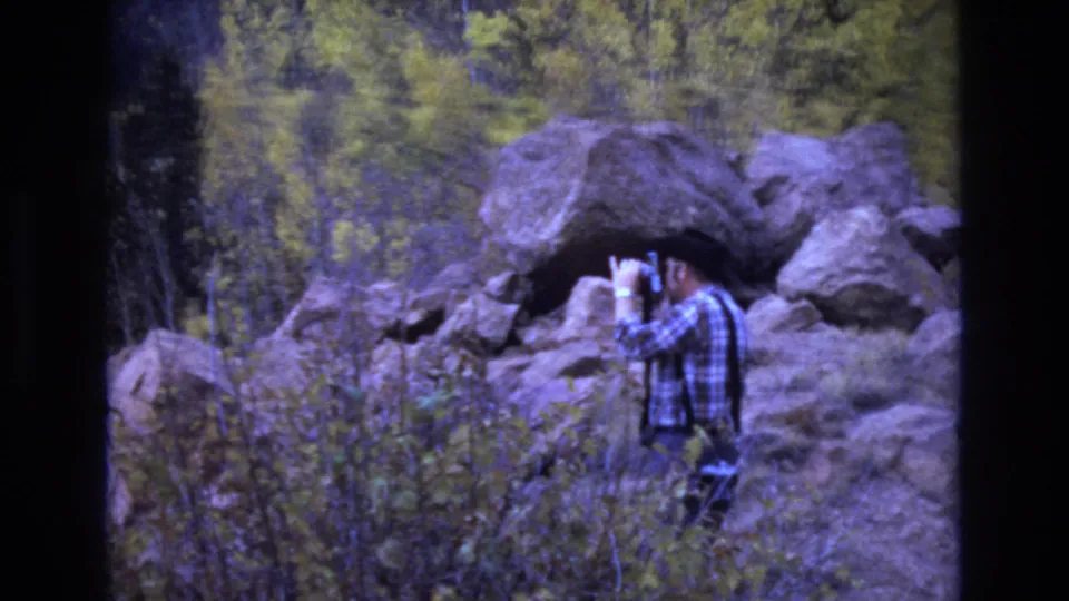 A man in a plaid shirt and dark hat holds a camera to his face as if taking a photograph in the rocky Colorado Springs forest setting.